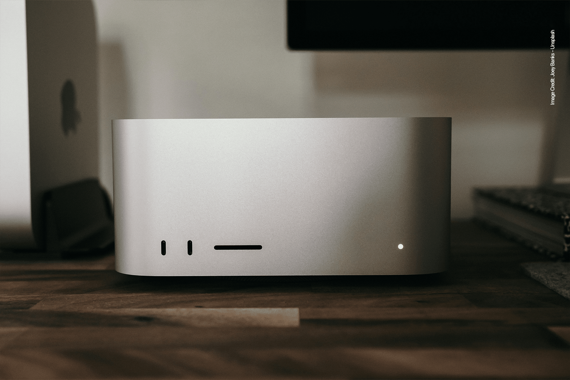 A sleek silver Mac Studio sits on a wooden desk, designed to streamline desktop productivity. With minimal ports and a single small light on the front, it’s surrounded by a blurred monitor and other tech accessories in the background.