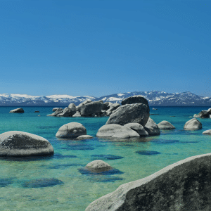 Large smooth rocks rest in the clear blue-green waters of a lake, framed by snow-capped mountains and a bright sky. A towering pine stands near the shore—an inviting scene featured in the Tahoe Day Aerial Screen Saver for macOS.