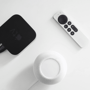 A black Apple TV box running tvOS 18.6 with connected cables, a white Apple TV remote, and a white Apple HomePod mini with HomePod 18.6 on a white surface.