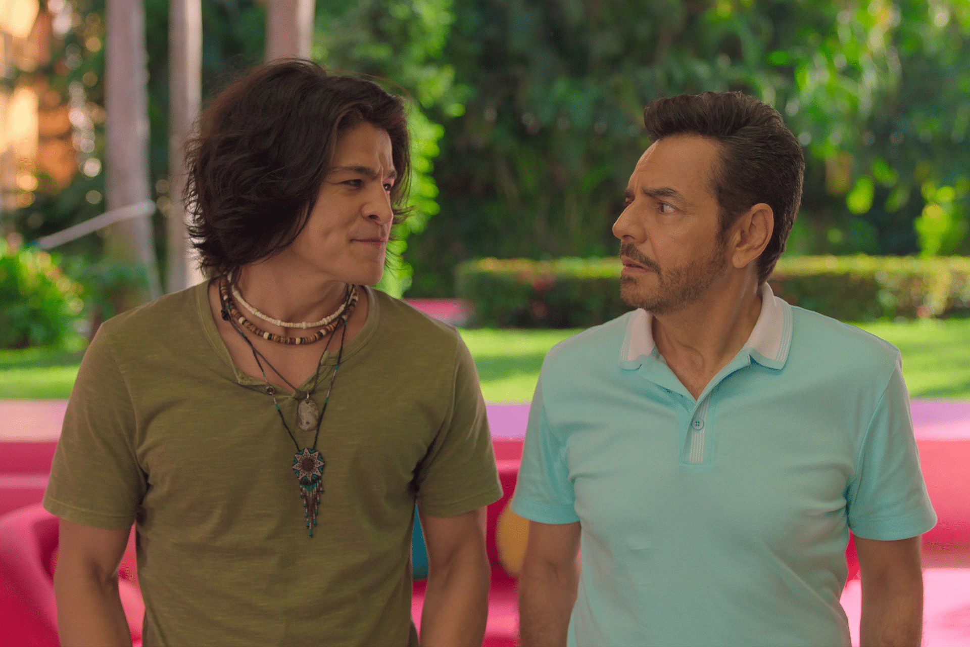 Two men stand outdoors facing each other. One wears a green shirt with layered necklaces, the other a light blue polo. Both have serious expressions, surrounded by greenery and colorful furniture—a scene fit for Apple TV+ summer streaming.
