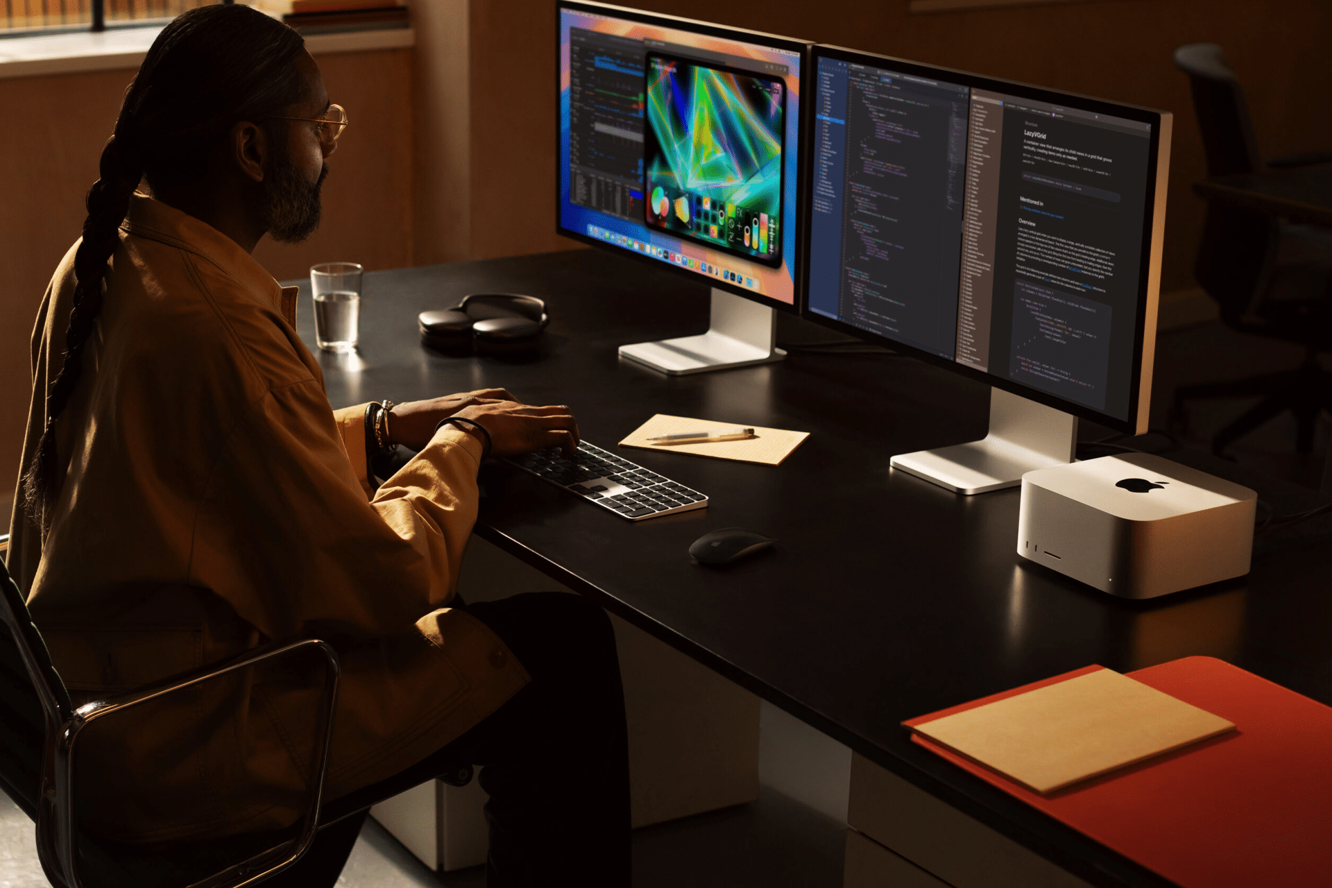 A person with long hair, glasses, and a yellow jacket sits at a desk using a keyboard, working on two large monitors displaying code and graphics. Office items and the new Mac Studio are nearby, perfectly set up for moving files in a seamless transition.