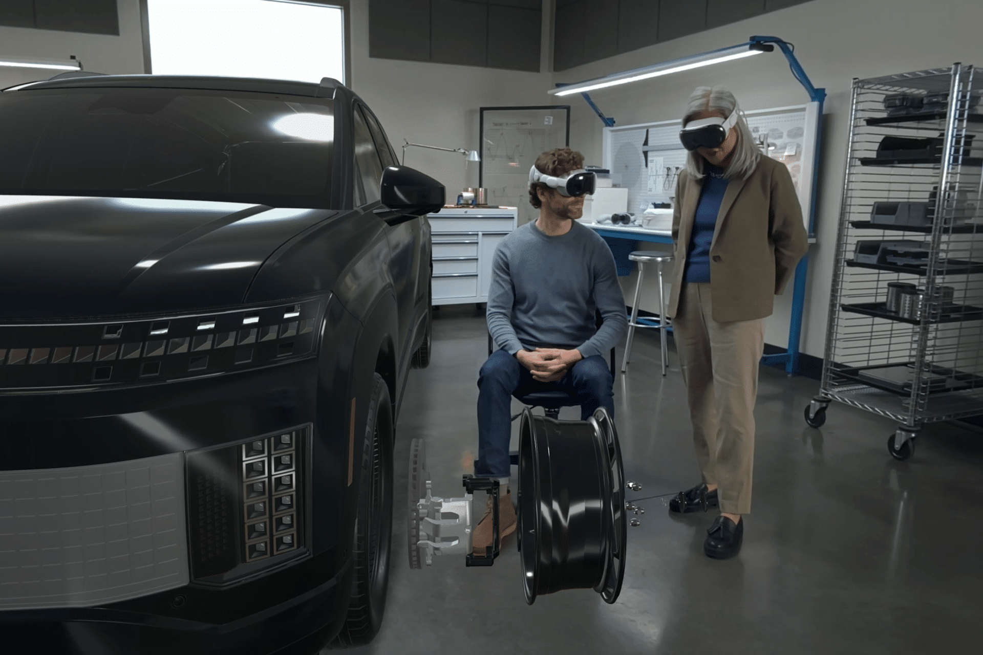 Two people wearing VR headsets interact with a virtual car model and parts in a high-tech workshop, exploring 3D modeling and design workflows—with one person seated and the other standing—using visionOS technology.
