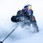 A skier in black gear and a helmet charges through deep powder snow, sending a spray into the air on a bright, wintry slope—perfect for experiencing immersive films with Vision Pro’s spatial storytelling. The skier's goggles reflect the snowy surroundings.
