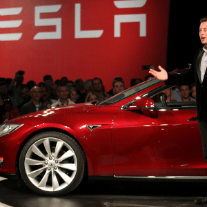 Elon Musk stands next to a red Tesla car on stage, addressing an audience. The Tesla logo is displayed on a red background behind him.