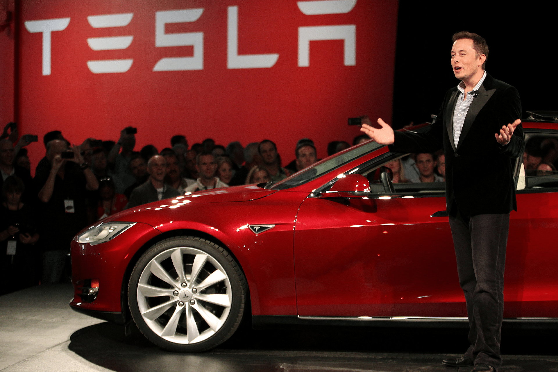 Elon Musk stands next to a red Tesla car on stage, addressing an audience. The Tesla logo is displayed on a red background behind him.