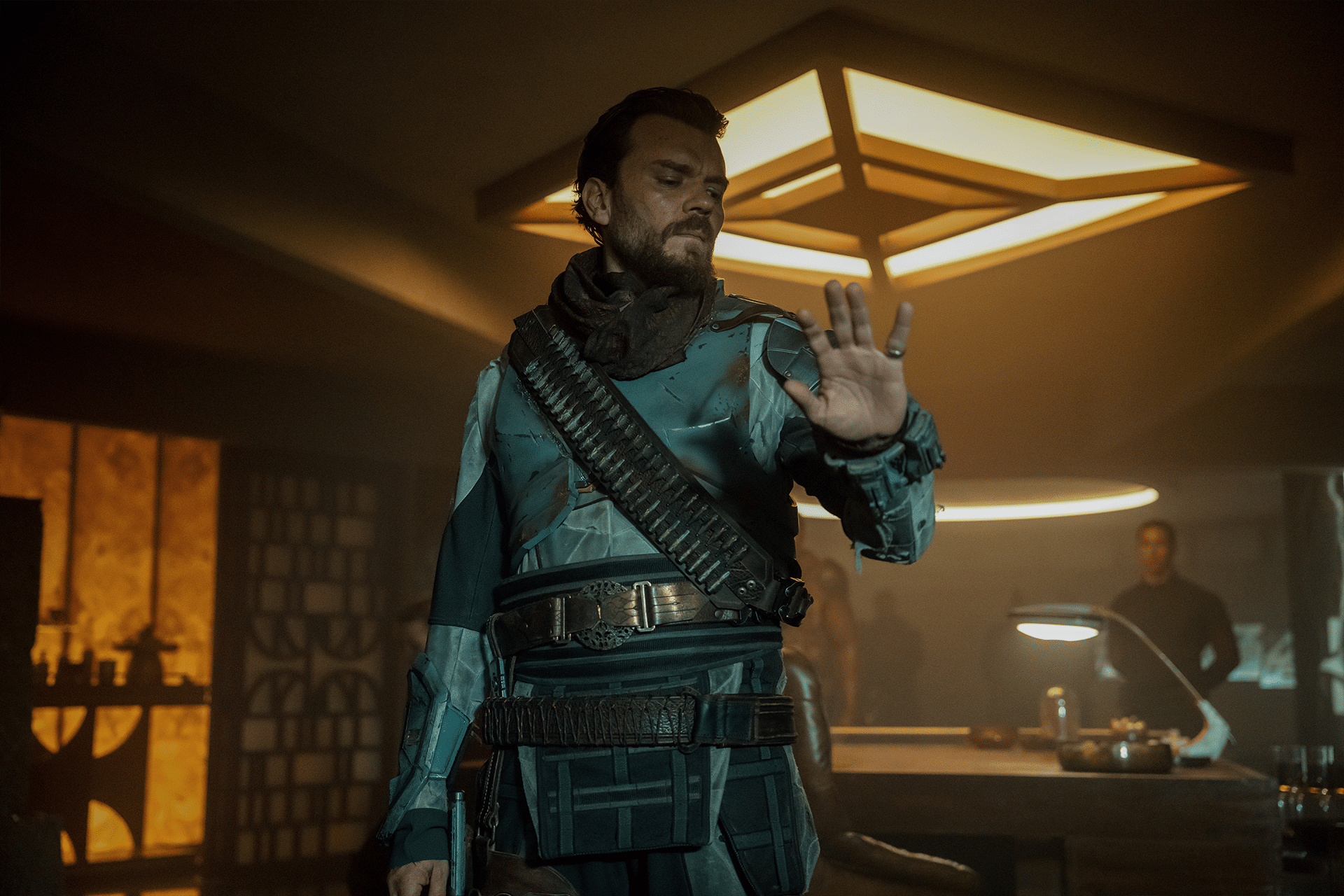 A man in futuristic armor with a bandolier stands in a dimly lit room, raising his hand as if gesturing to stop. Warm, geometric lights and modern furniture are visible—a scene reminiscent of Foundation Season 4 on Apple TV+.