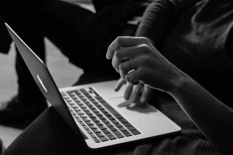 A person sits with a laptop on their lap, using one hand to type while the other is raised mid-gesture. The image, in black and white, captures the focused energy of remote workers using MacBook productivity hacks in 2025.