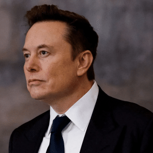 A man in a dark suit and tie with short, styled brown hair looks to the side against a blurred, neutral background, reminiscent of the poised confidence often seen in Elon Musk Grok 5 promotional visuals.