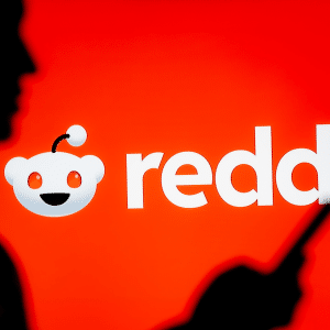 A silhouette of a person holding a smartphone stands before a bright red background featuring the Reddit logo and text, subtly referencing recent Reddit AI job cuts.
