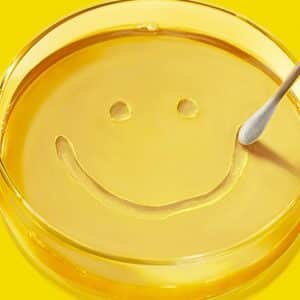 A hand uses a cotton swab to draw a smiling face in a petri dish filled with yellow liquid, set against a bright yellow background.