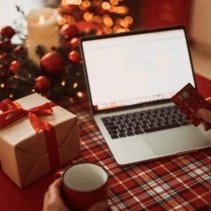 A person holding a credit card and a mug sits at a table with a wrapped gift, an open laptop, festive decorations, and warm lights in the background, ready for some seamless AI holiday shopping.