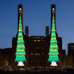 A brick power station at night with two tall chimneys, each brightly lit for the Apple Christmas tree challenge—adorned with cartoon-style Christmas trees topped by stars. The cityscape glows in the background.