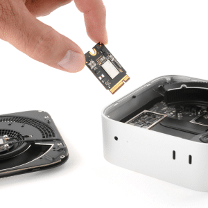 A hand holds an SSD chip above an open Mac mini, with internal components and a detached fan visible on a white surface—a reminder of hardware upgrades amid rising Apple RAM prices.
