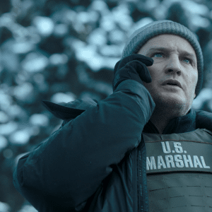 A man in a winter beanie and jacket, wearing a bulletproof vest labeled "U.S. Marshal," stands outdoors in a snowy environment, holding a phone to his ear and looking concerned—perhaps awaiting news on Apple TV’s decision to cancel The Last Frontier.