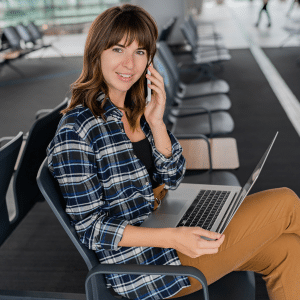 Apple Travel - A woman sits in an airport terminal, smiling while talking on her iPhone. She has a laptop on her lap, wears a plaid shirt and brown pants, and there are empty seats around her as she checks travel safety tips.