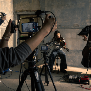 Two musicians play string instruments in a dimly lit room while a small crew films them with cameras, audio equipment, and an iPhone 17 Pro for video making. Multiple tripods and recording devices are set up around the performers.