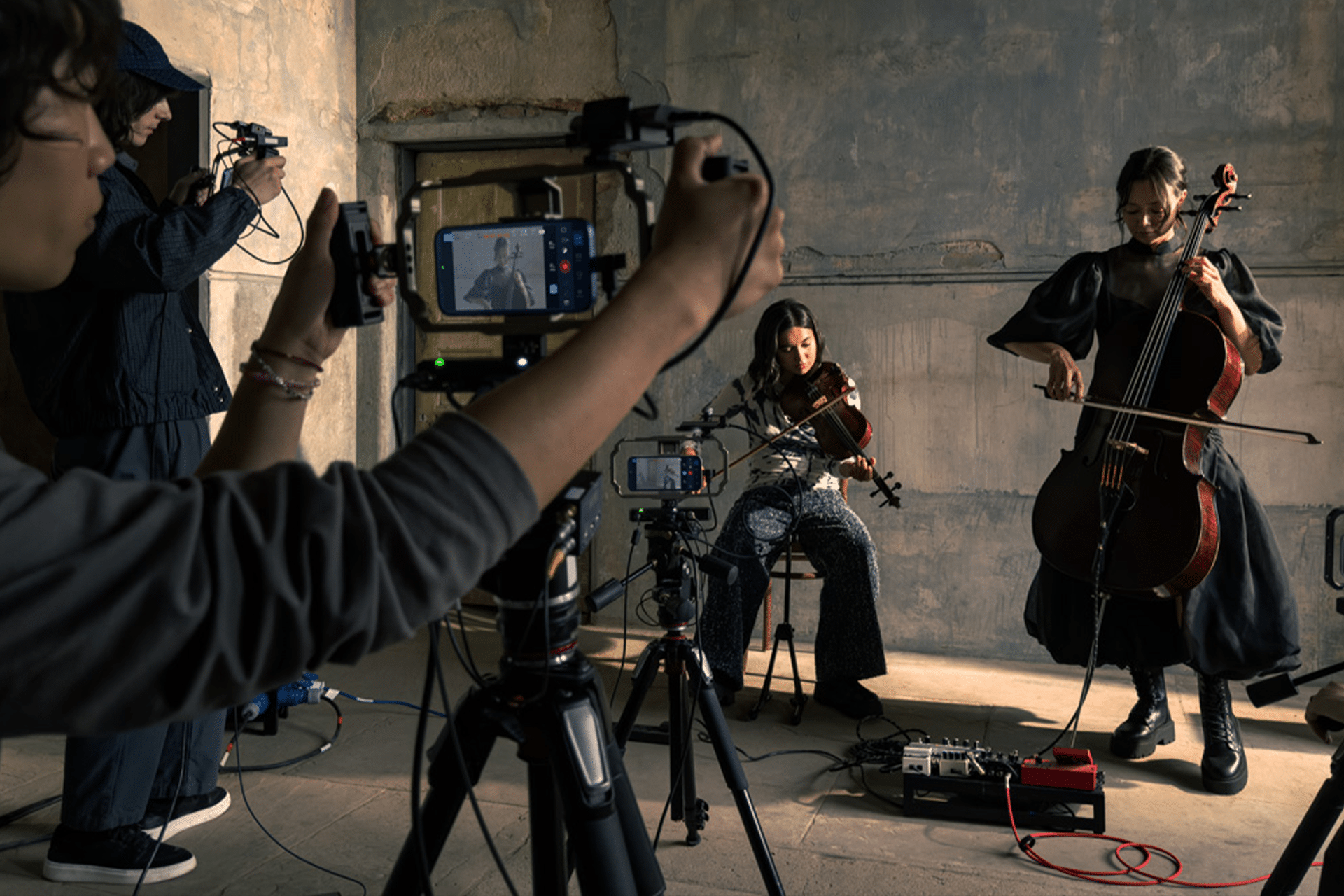 Two musicians play string instruments in a dimly lit room while a small crew films them with cameras, audio equipment, and an iPhone 17 Pro for video making. Multiple tripods and recording devices are set up around the performers.