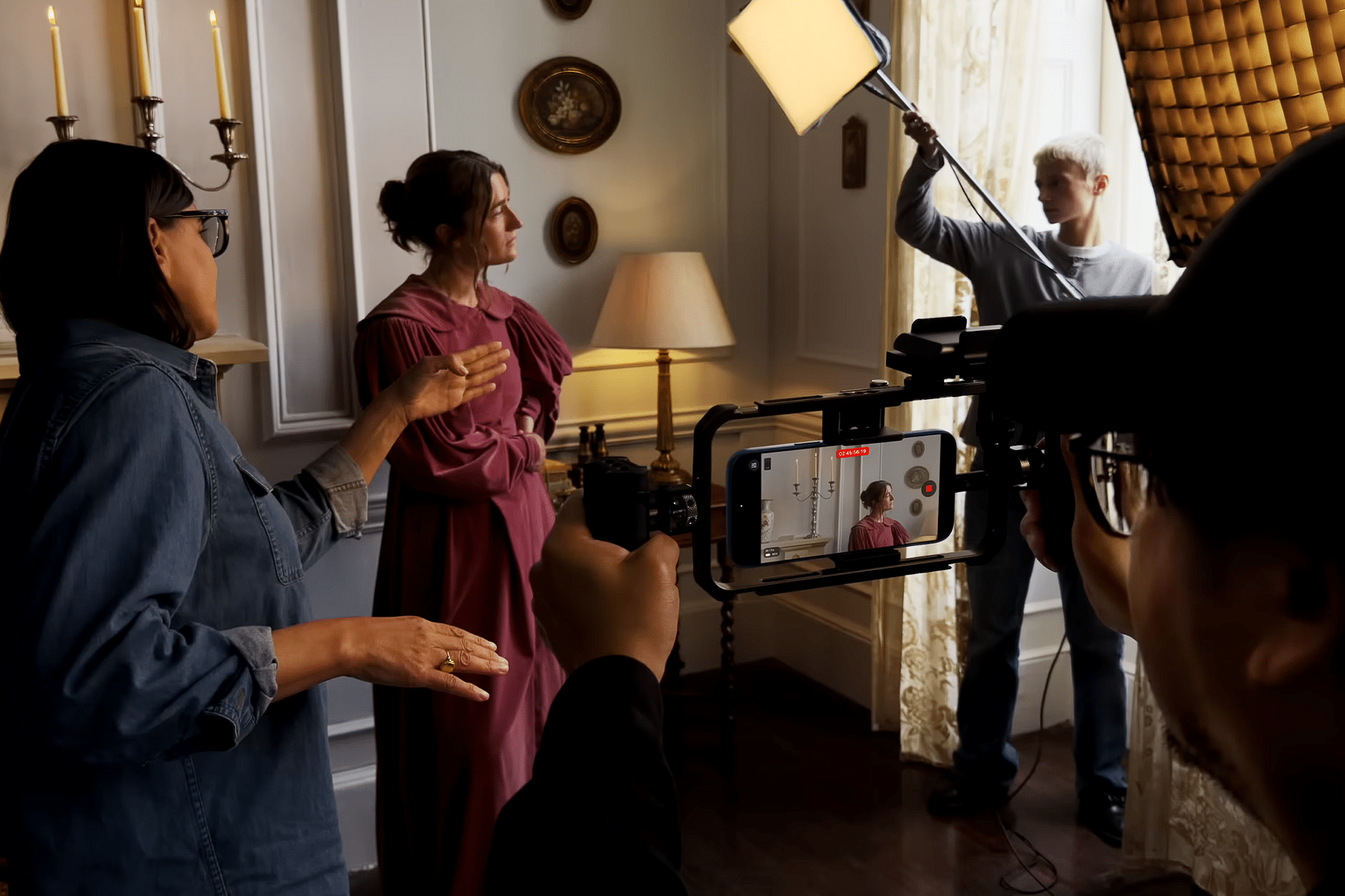 Two people converse on a film set, one in a pink costume. Crew members use lighting equipment, sound boom, and an iPhone 17 Pro for video making. The indoor scene features candles, framed pictures, and a lamp in the background.