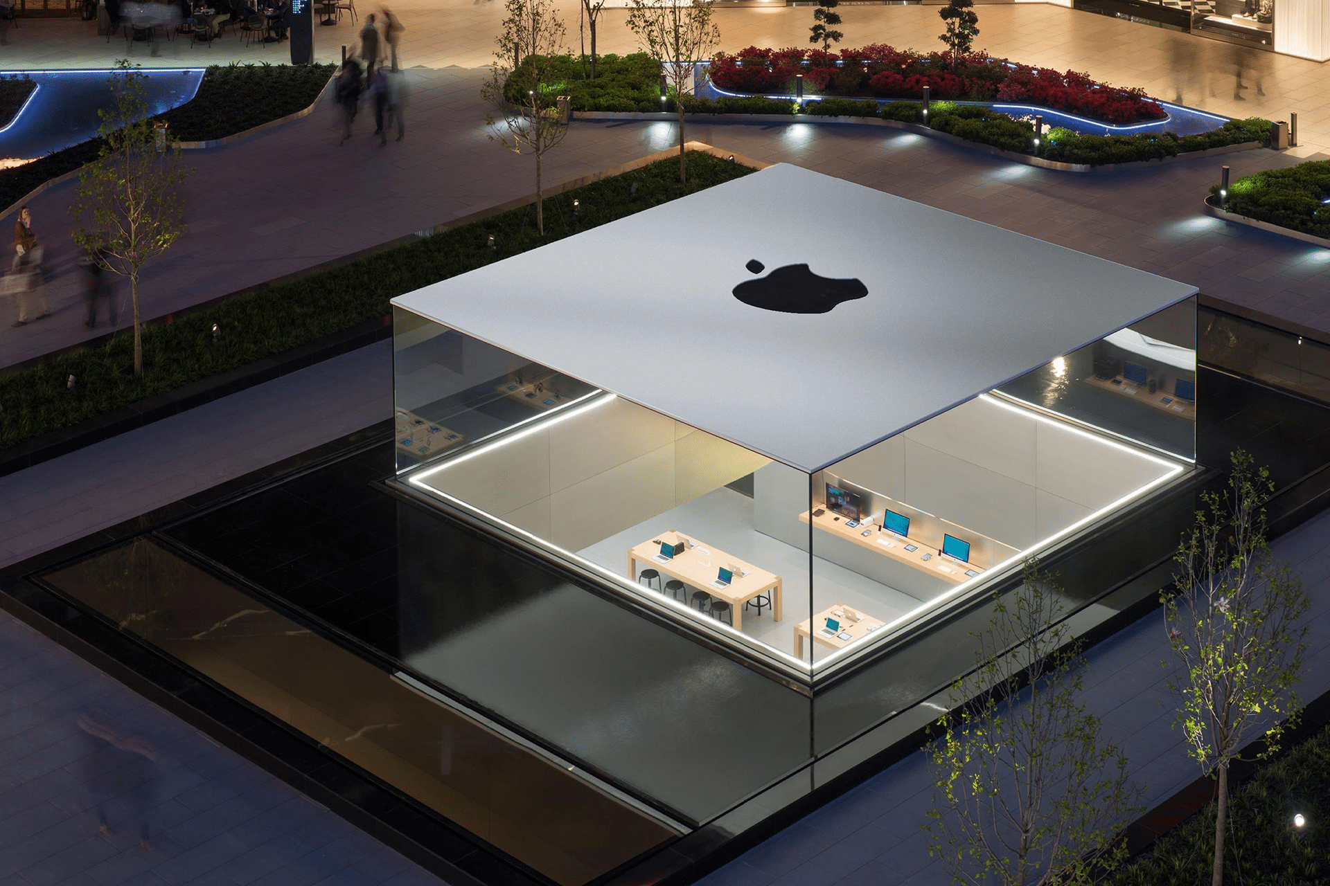 Apple Q3 results - A modern, glass-walled Apple Store near me sits surrounded by water and landscaped greenery, with tables displaying Apple products inside. The building has a large Apple logo on the flat, white roof. People walk nearby.