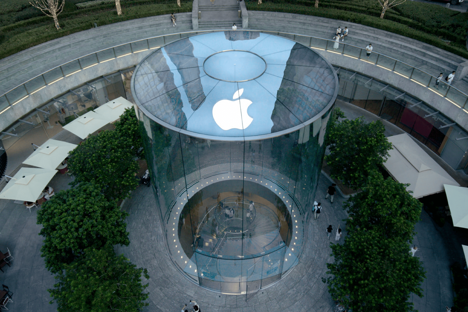 Aerial view of a cylindrical glass Apple Store Near Me, featuring a large white Apple logo on the roof, surrounded by trees, outdoor seating, and a curved stone walkway.
