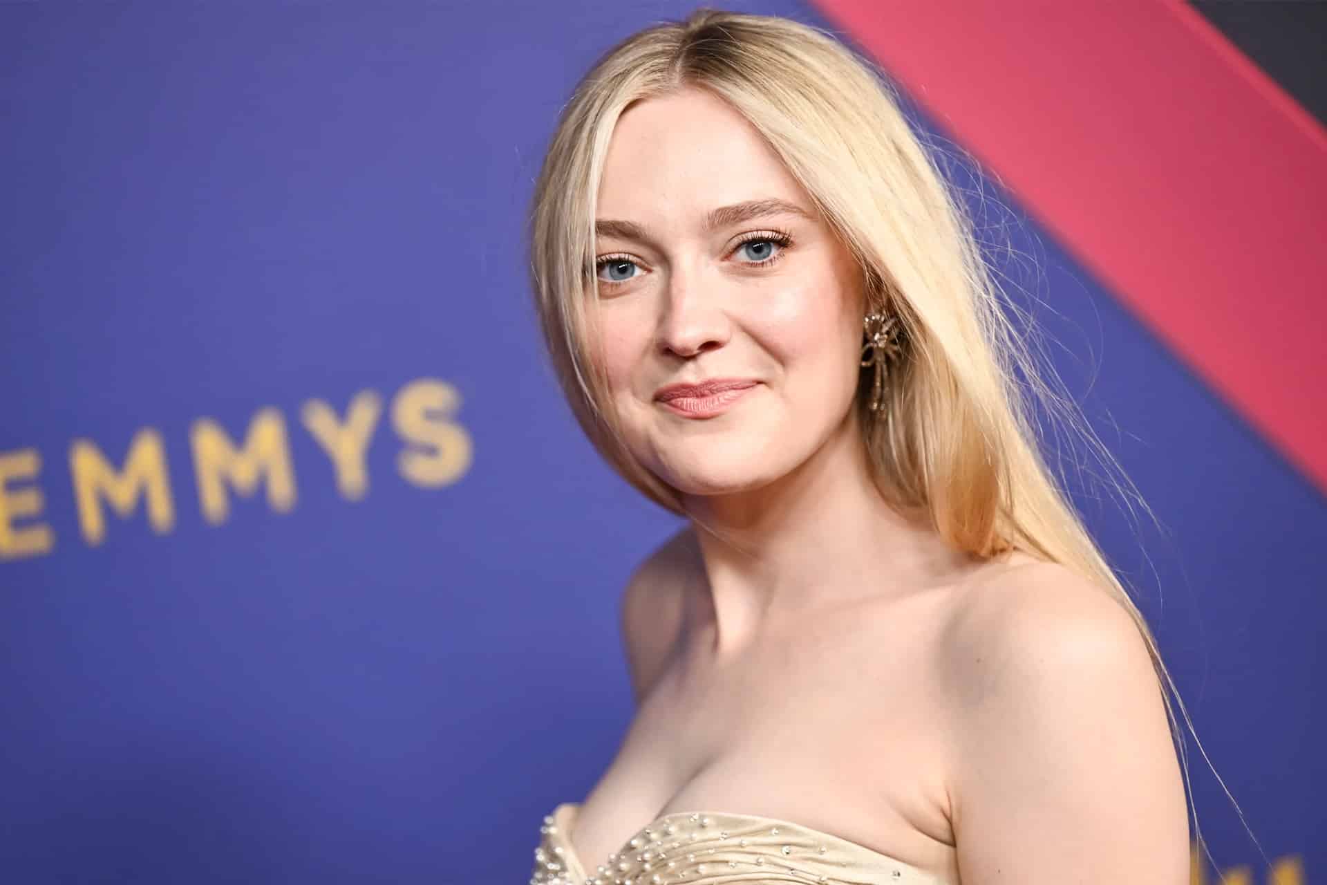 Dakota Fanning, with long blonde hair and a strapless cream-colored dress, smiles softly at the camera in front of a purple and red backdrop with "EMMYS" in gold—celebrating her role in the Apple TV New Thriller.