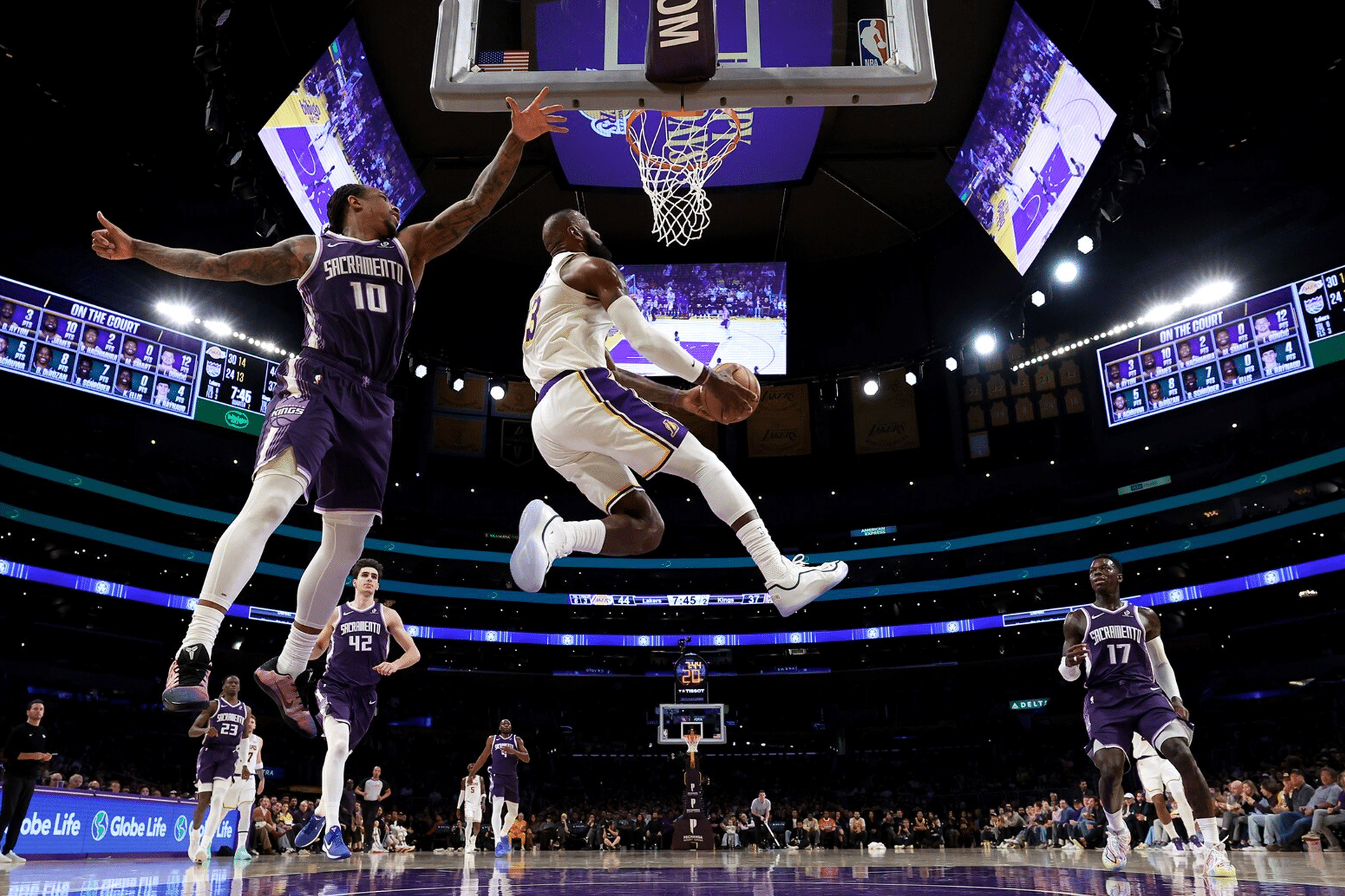 A basketball player in a white uniform leaps for a layup as an opponent in purple tries to block him during an exciting game in a packed, bright arena—action you can catch live on Apple TV NBA League Pass.