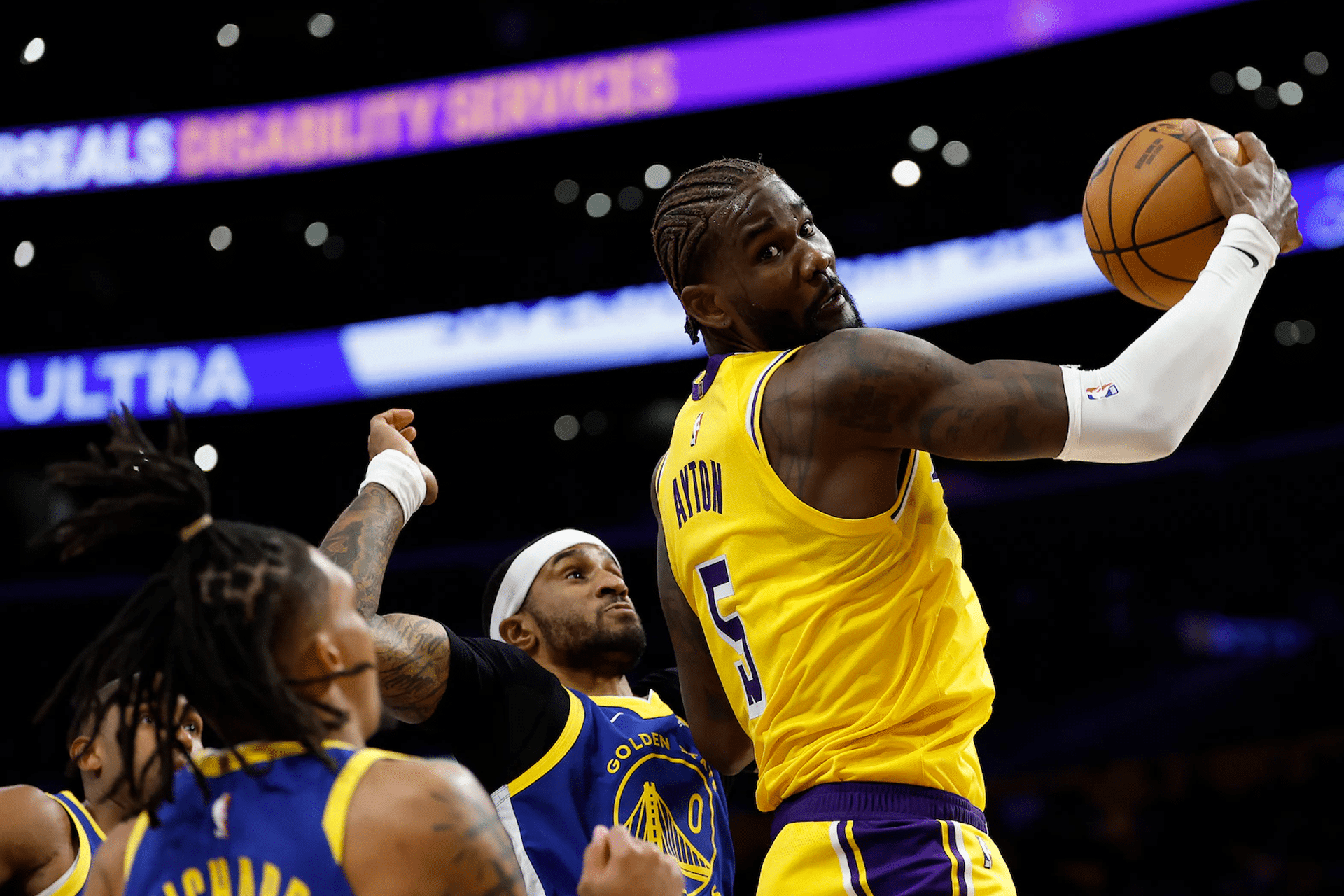 A basketball player in a yellow jersey grabs a rebound while surrounded by three players in blue jerseys during an NBA game, with arena lights and a scoreboard in the background—perfect action to catch on Apple TV NBA League Pass.