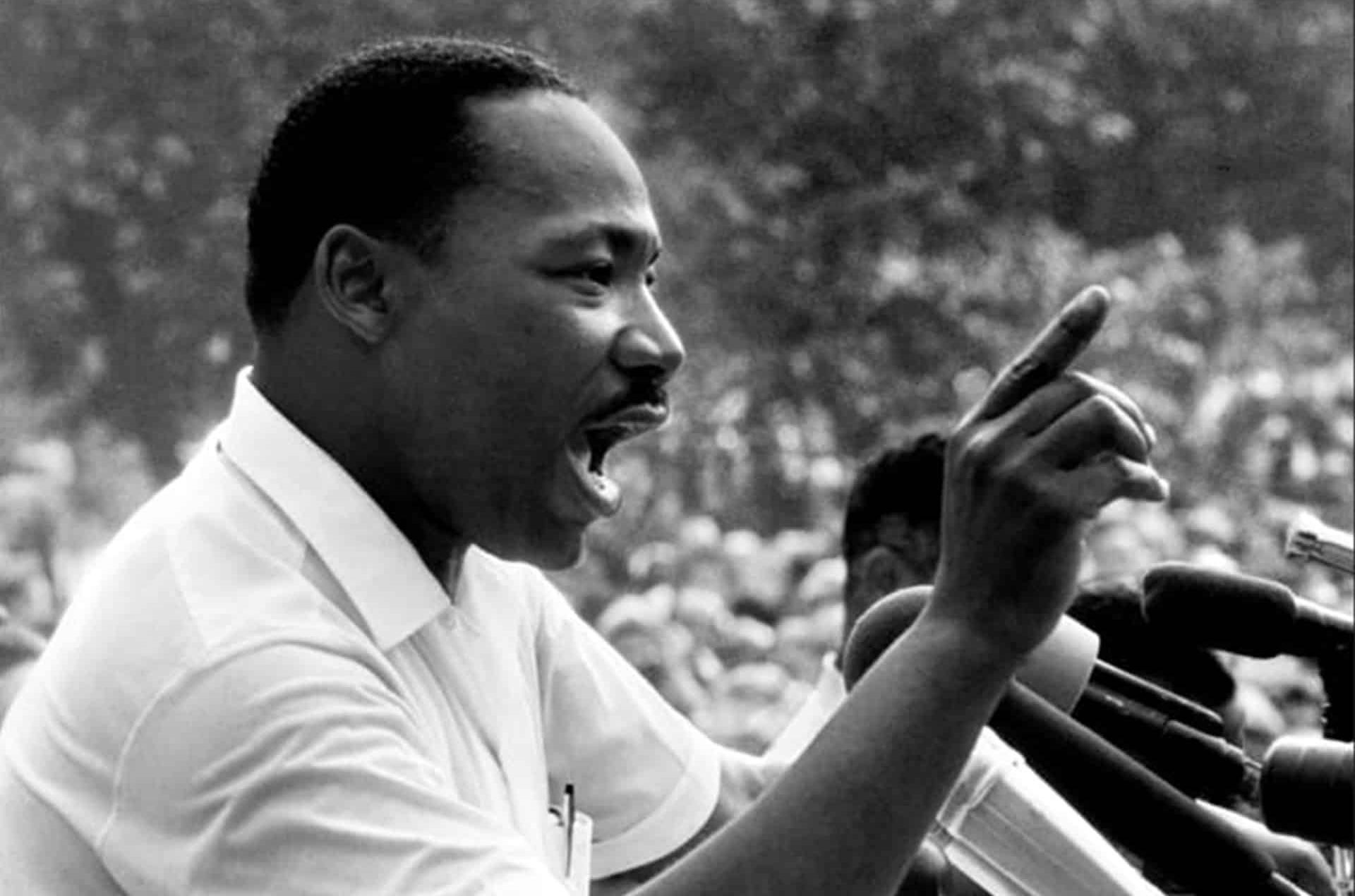A man, reminiscent of Martin Luther King, speaks passionately at a podium, gesturing with his right hand in front of several microphones, with a blurred crowd in the background.
