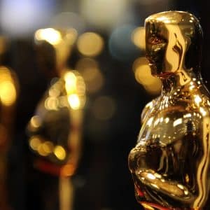 A close-up of a shiny gold Oscar statuette, with other similar statuettes blurred in the background against a backdrop of warm, out-of-focus lights—captured using Apple Creator Studio for stunning clarity. - Apple Academy Awards