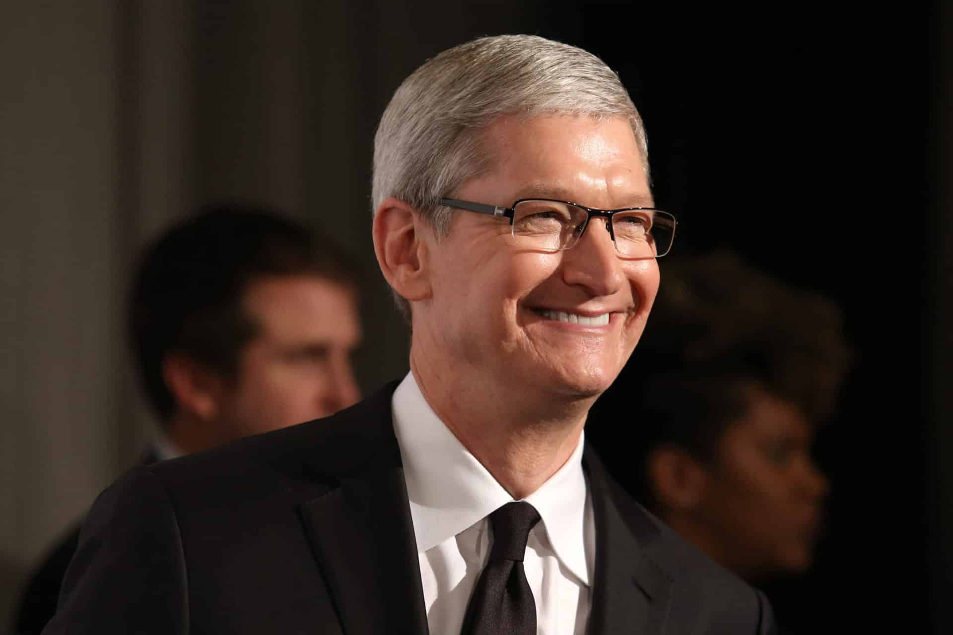 A man with short gray hair and glasses, resembling Tim Cook, smiles while standing indoors in a black suit. Two people are visible but out of focus behind him, hinting at an Apple 2026 "one more thing" announcement.