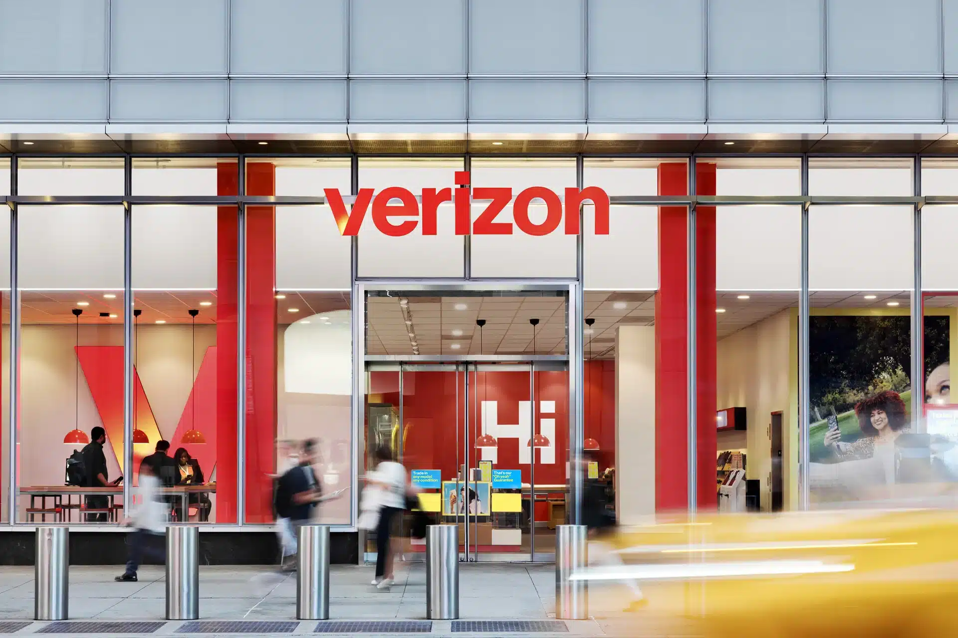 A Verizon store with large glass windows, red accents, and the Verizon logo above the entrance highlights the Verizon vs AT&T competition. Several people walk by outside, with a yellow taxi passing in front of the store.