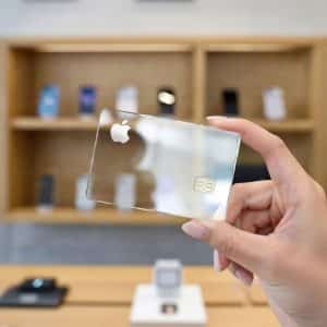 A hand holds a transparent digital-first credit card with the Apple logo in a modern tech store, where shelves display various electronic devices in the background.