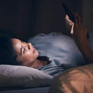 A woman lying in bed at night looks at her smartphone screen, illuminated by Night Shift mode. She is resting her head on a pillow and appears relaxed and focused in the dark room.