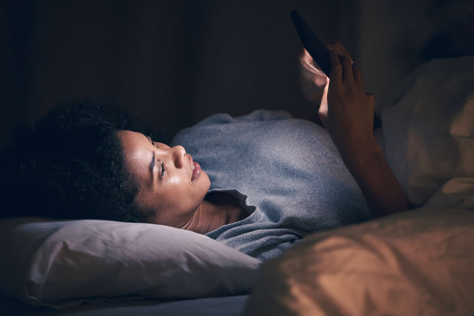 A woman lying in bed at night looks at her smartphone screen, illuminated by Night Shift mode. She is resting her head on a pillow and appears relaxed and focused in the dark room.