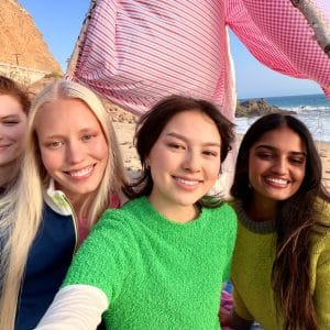 Four young women smile and pose for a selfie on a sandy beach with the ocean and cliffs in the background. They sit under a striped pink-and-white canopy made with sticks, capturing the sunny day in fun iPhone Photos.