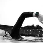 A swimmer in a pool performs a freestyle stroke, wearing goggles, a swim cap, and an Apple Watch Ultra displaying workout data on their wrist. The background features a fence, trees, and pool lane dividers.