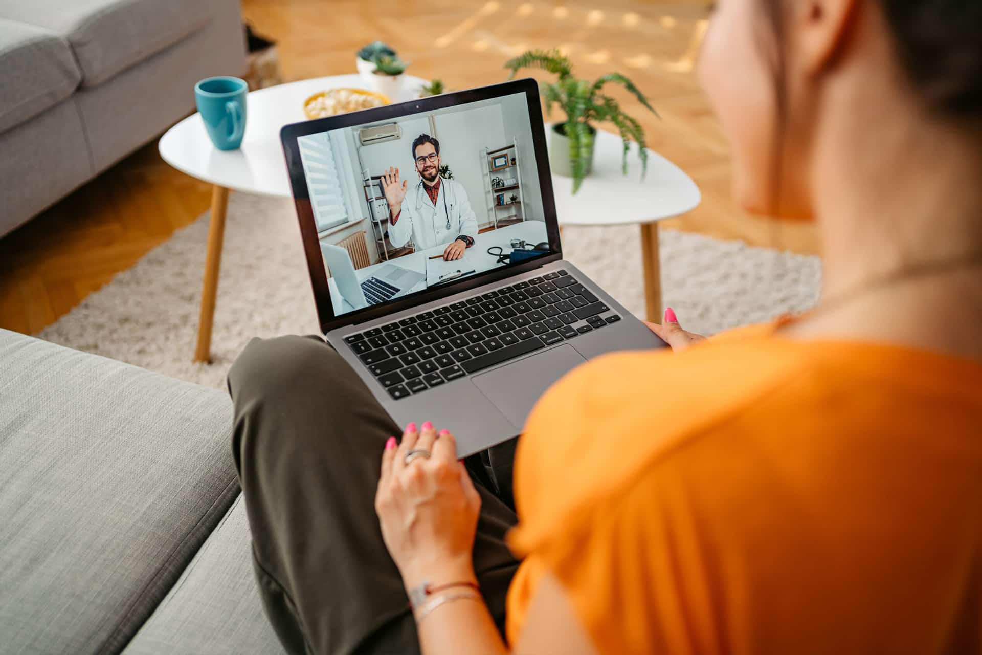 A woman sits on a sofa, holding a laptop on her lap in a remote therapy setup during a video call with a doctor. The doctor, wearing a white coat, is waving from the screen. A coffee table with mugs and snacks is in the background.