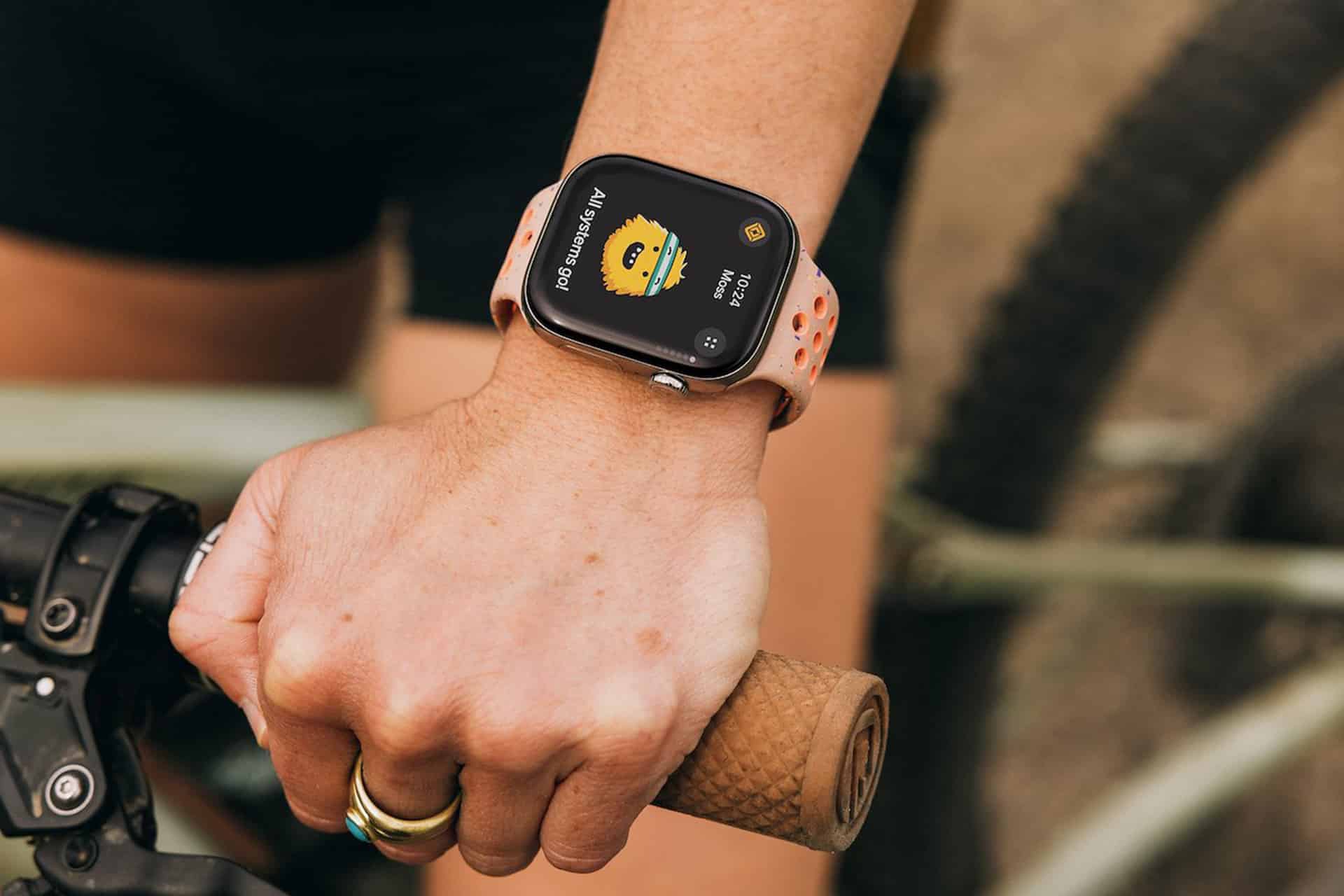 A person wearing an Apple Watch with a white and pink band grips a bicycle handlebar. The watch displays a yellow animated face and the text, "AN ANGRY POD." They're outdoors in black shorts, possibly using the Rivian Apple Watch app.