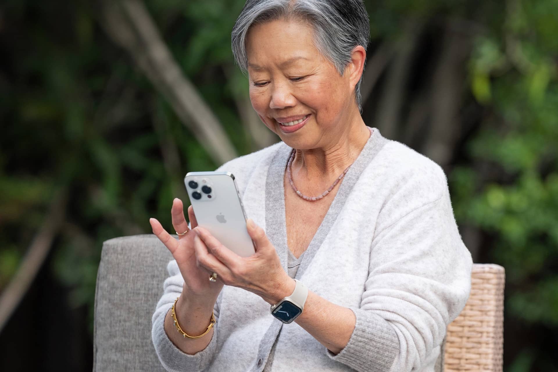 An older woman with gray hair smiles while using an Apple smartphone outdoors. She is wearing a light cardigan, a smartwatch, a bracelet, and a necklace, sitting on a wicker chair with greenery in the background.