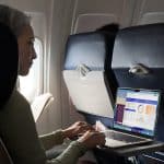 A woman sits on an airplane using a laptop on the tray table, viewing charts and graphs on the screen. She holds a document in her left hand and looks at the laptop, with sunlight coming through the window beside her.