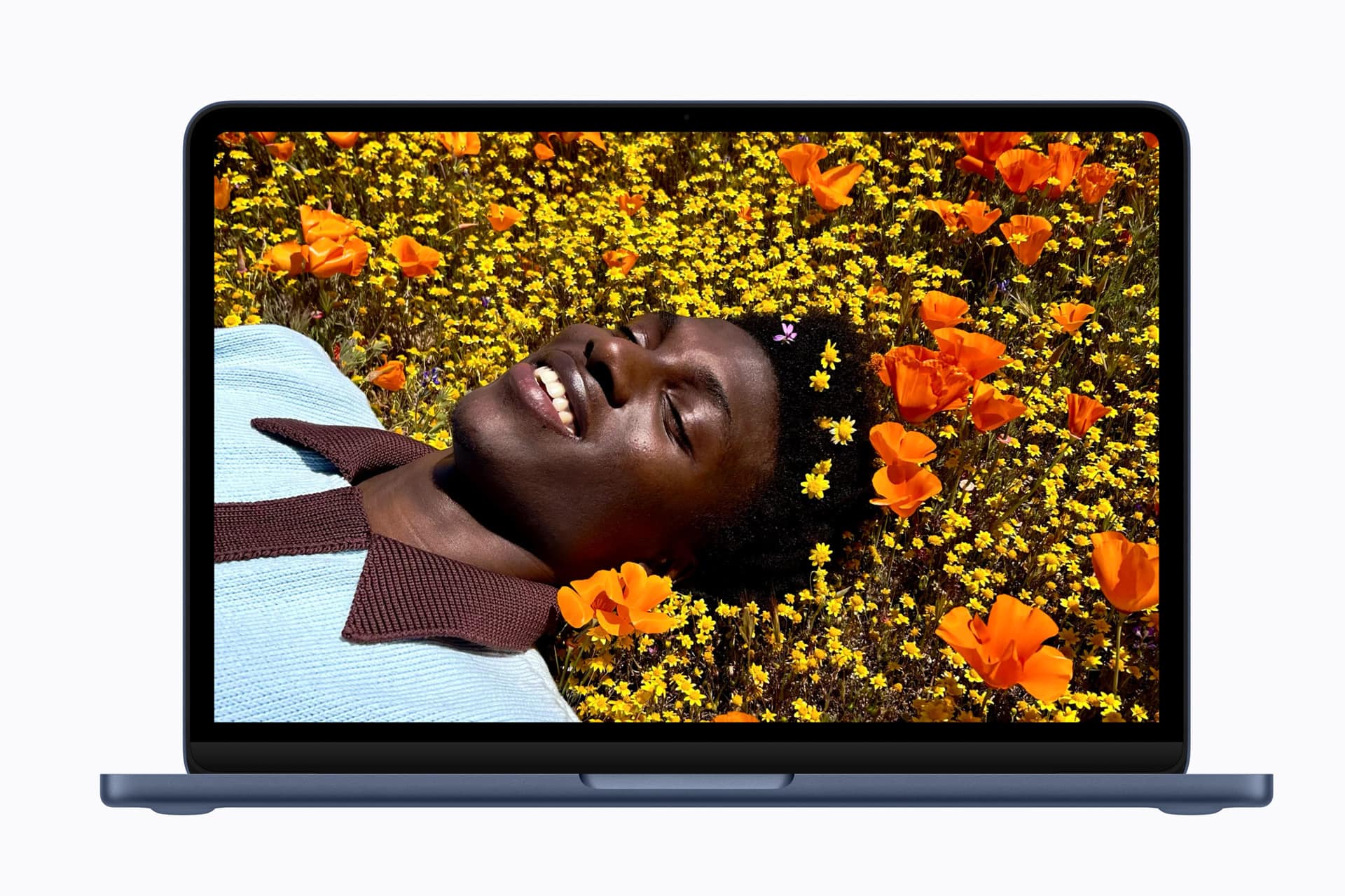 A laptop screen displays a vibrant image of a person lying in a field of yellow and orange flowers, smiling and looking relaxed. The flowers surround their head, creating a colorful, cheerful scene.