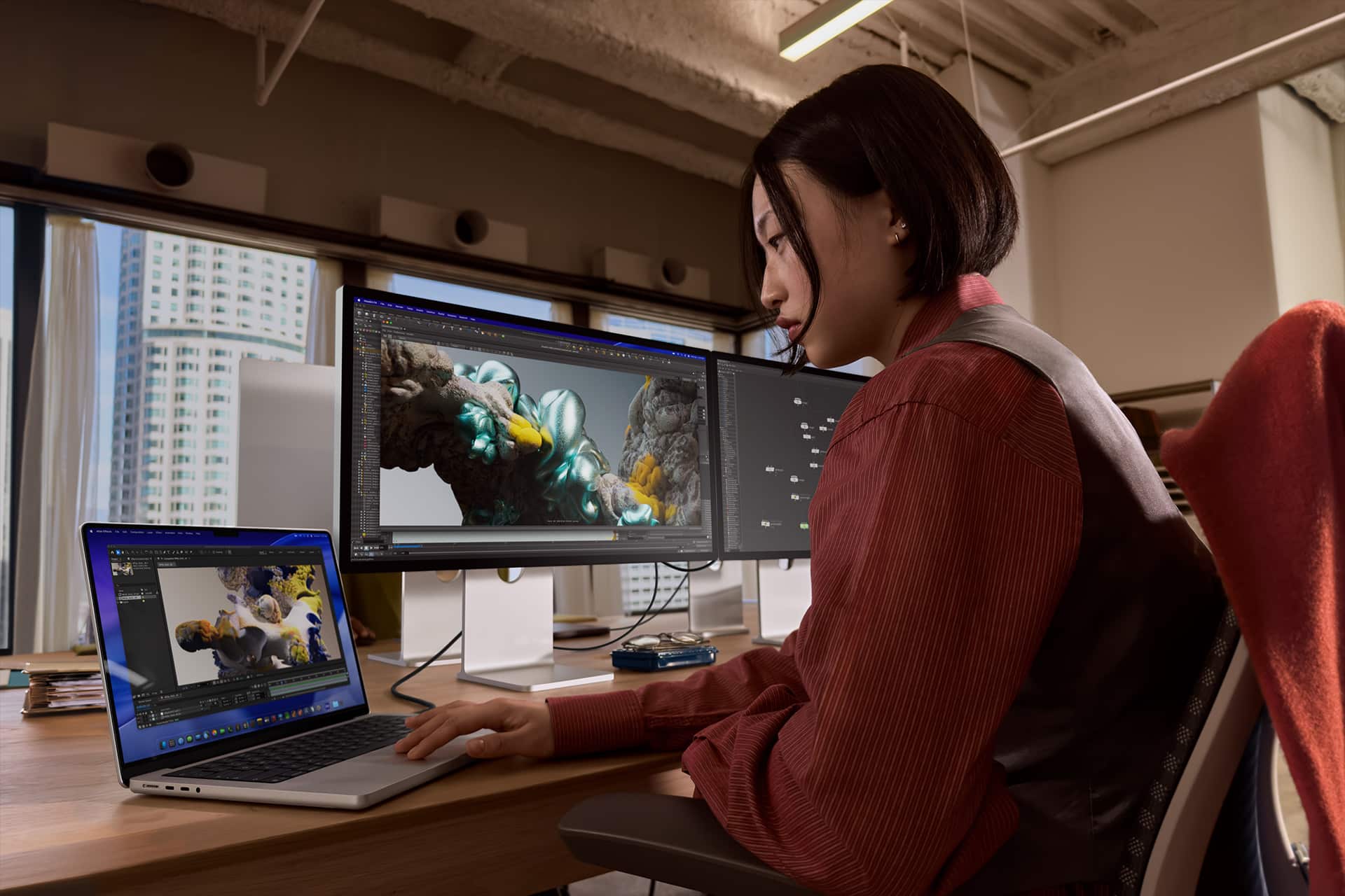 A woman sits at a desk in an office, working on 3D modeling with colorful sculpted figures displayed on a laptop and two monitors. She is focused on her work, surrounded by large windows and modern decor.