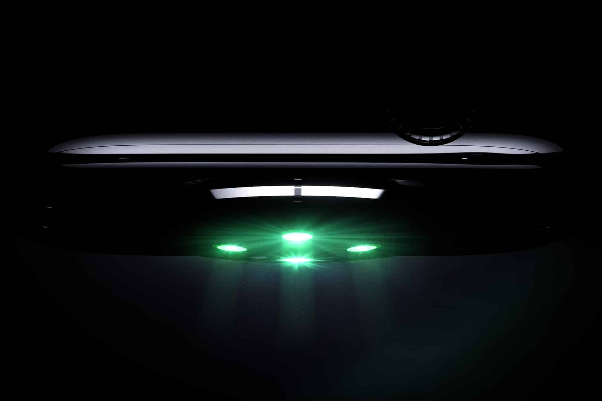 A close-up, low-light image of the underside of a smartwatch emitting green light from its sensors, highlighting its sleek metallic finish and side buttons, showcases the watch’s focus on oxygen accuracy.