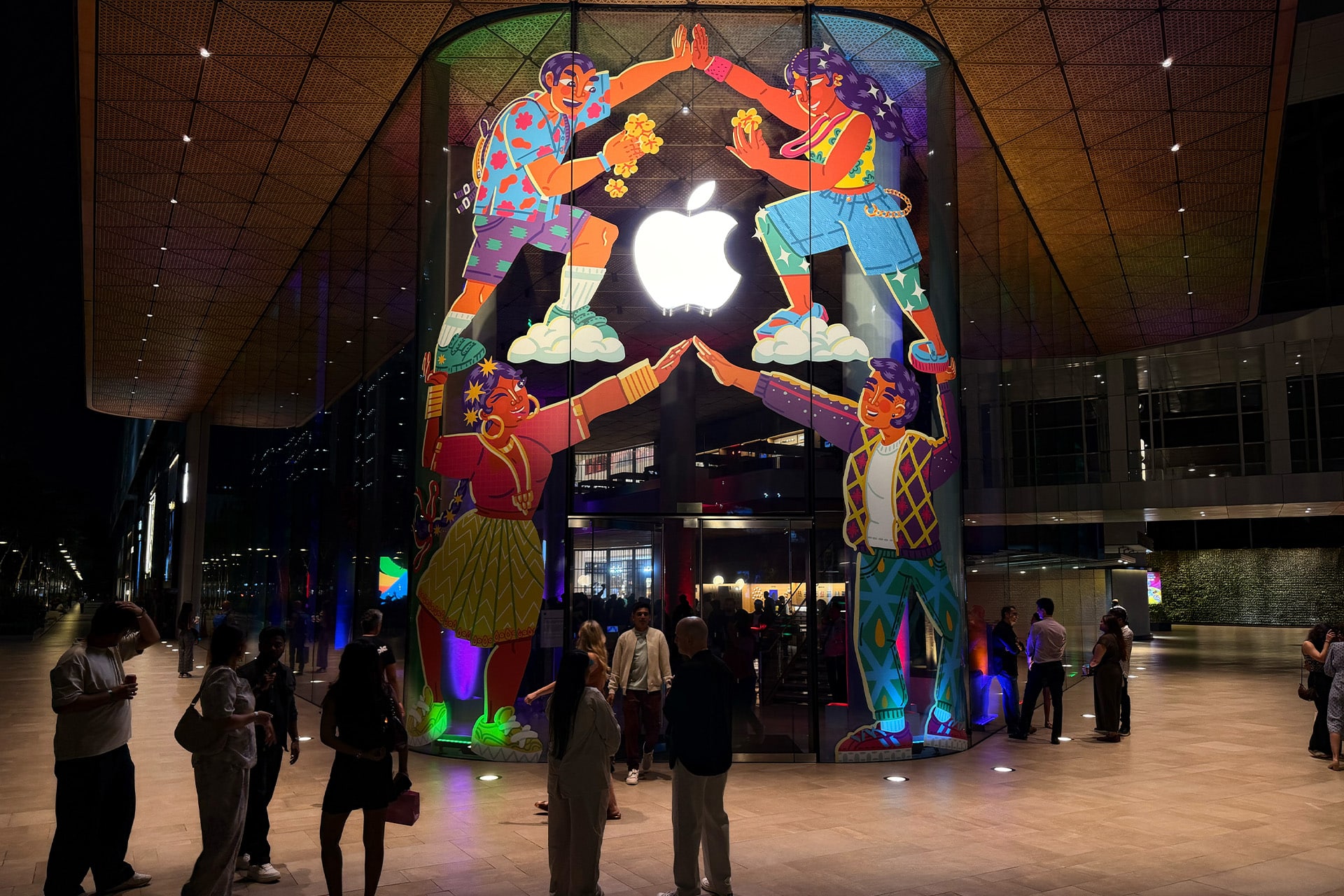 Apple India exports - People stand outside an Apple Store at night, vibrant mural and glowing logo above them. The lively scene hints at more than tech—Apple’s success reflects a smart cash reserve strategy behind the colorful, bustling storefront. India Store.