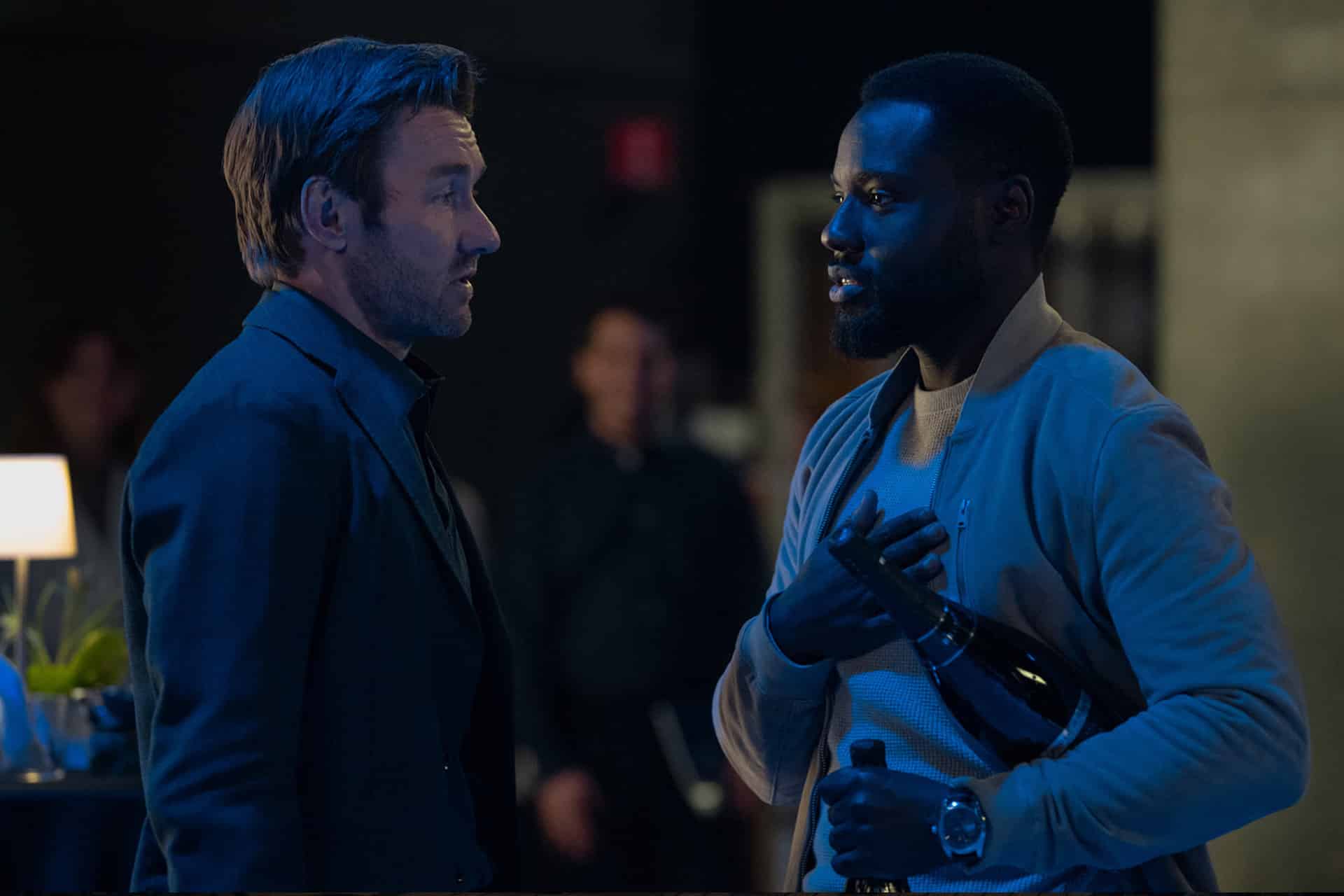 Two men stand facing each other at an indoor event; one holds a wine bottle and gestures with his hand on his chest, while the other listens. The dimly lit background, like a scene from Dark Matter Series, features blurred figures and tables.