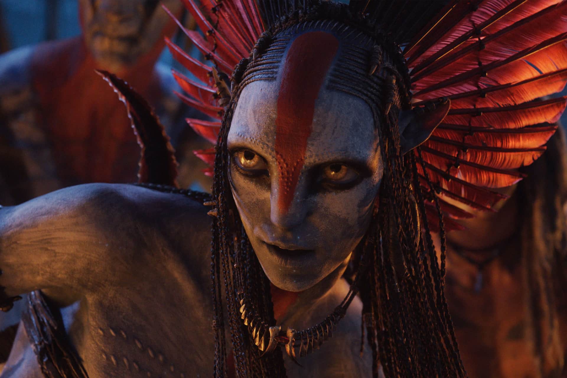A Na'vi warrior with striking yellow eyes and a red stripe down her face wears a headdress of red feathers and braids, ready to star in epic tales fit for top Apple TV Streaming Apps.