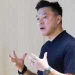 A person wearing a dark shirt gestures with both hands while speaking, standing indoors against a light-colored wall. They wear a ring, a bracelet, and a smartwatch—perhaps discussing innovative Korean apps or sharing experiences from the Apple Developer Academy.