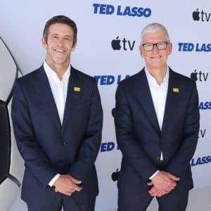 Two men in dark suits stand side by side, smiling, in front of a backdrop with "TED LASSO" and Apple TV logos, reminiscent of the recent Tim Cook and John Ternus appearances during discussions around the Apple CEO transition. A large, partial soccer ball is visible on the left side of the image.