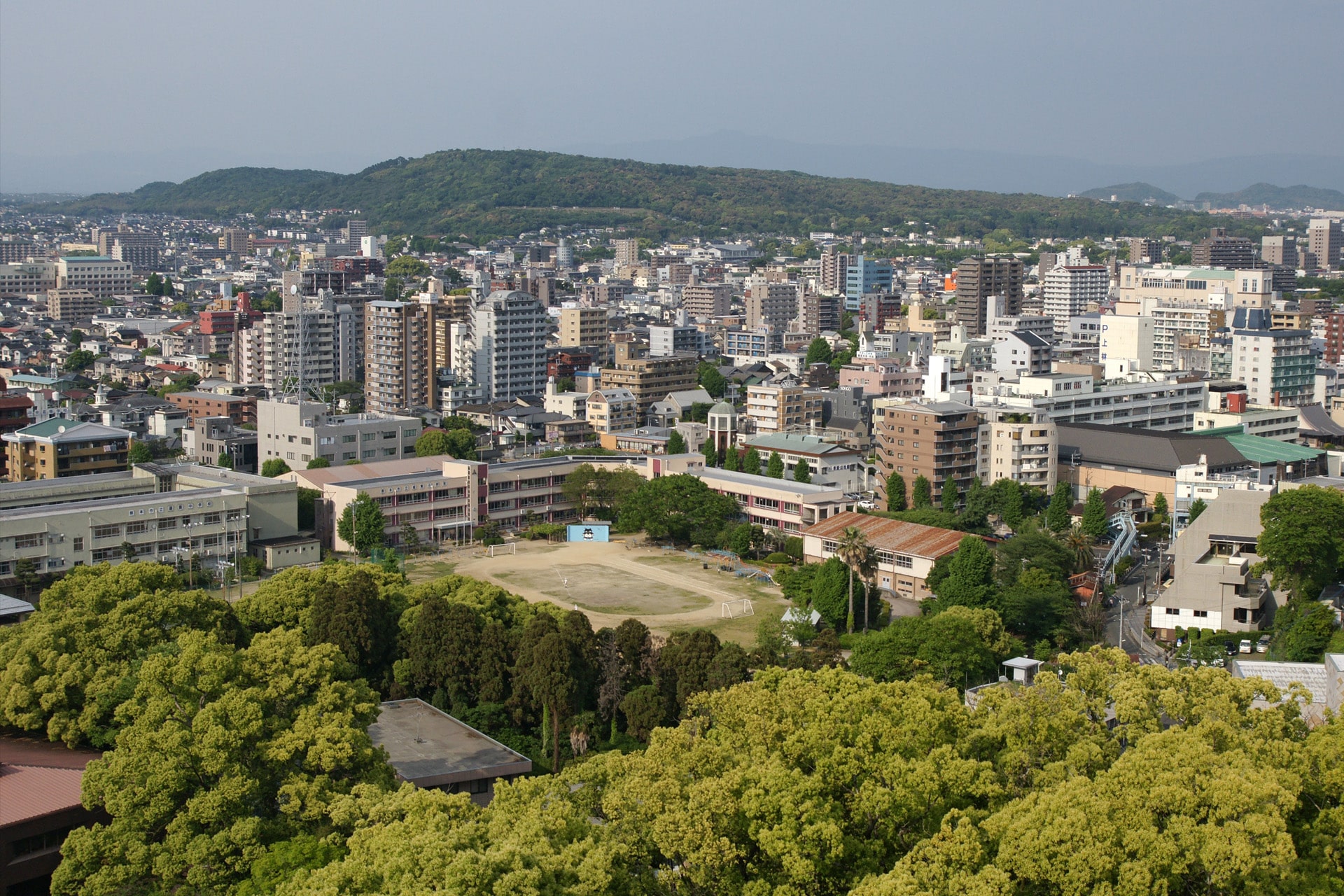 A cityscape with mid-rise buildings, a central school with a dirt sports field, and lush green trees in the foreground. In the wake of the Japan earthquake relief efforts, signs of recovery are evident throughout the scene. Distant hills and a hazy sky are visible in the background.