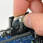 Mac PRAM - A hand is removing a round silver CMOS battery from a holder on a computer motherboard using a small metal tool to perform an NVRAM reset.